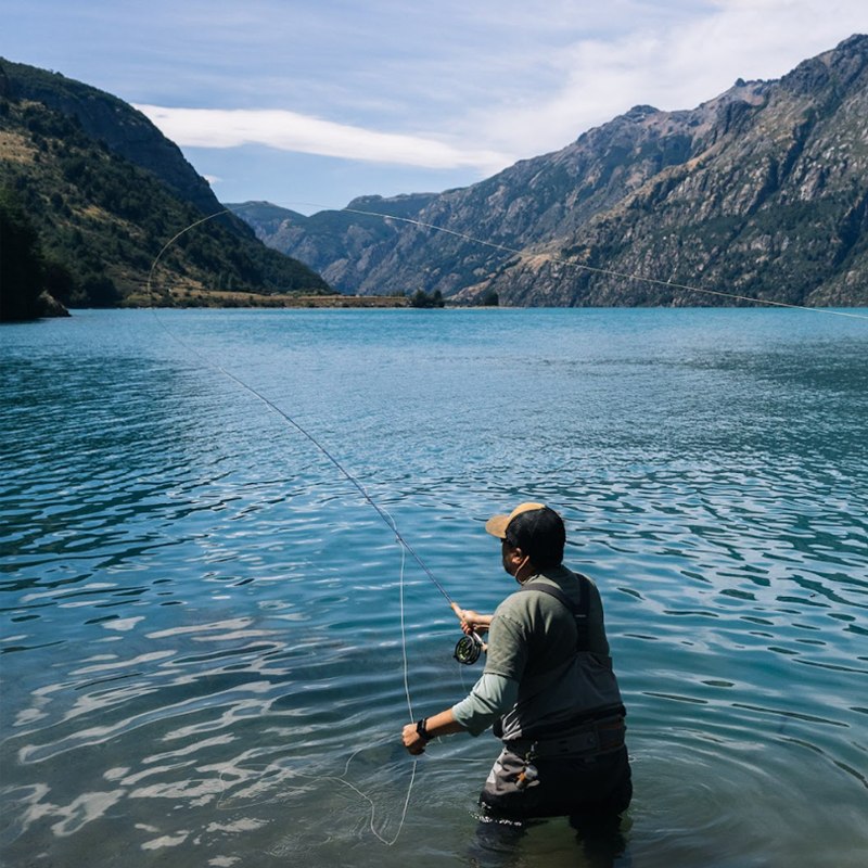 A man stands hip deep in a clear water lake surrounded by mountains on a lightly cloudy day