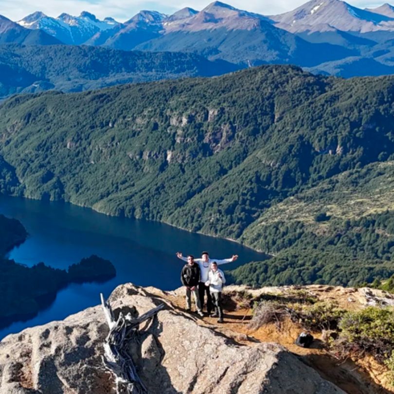 Three people with backpacks and trekking poles head up a grassy hillside to higher mountains.