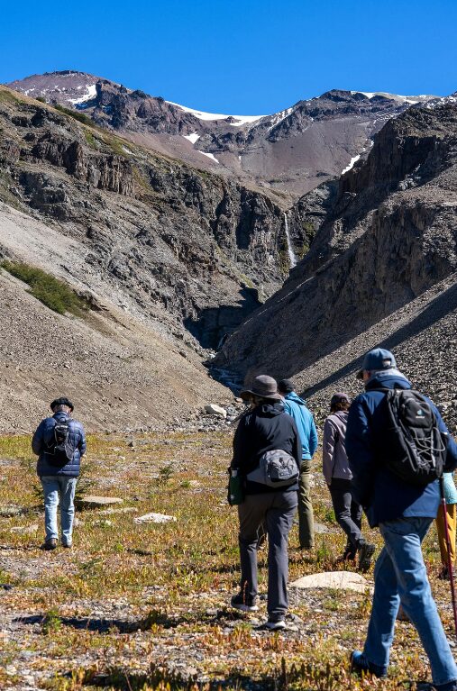 Three people with backpacks and trekking poles head up a grassy hillside to higher mountains.