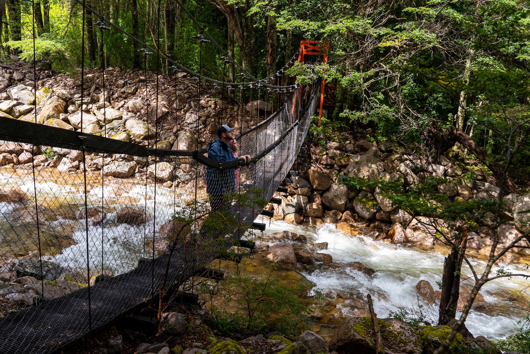Two people stand on a rope bridge over a river