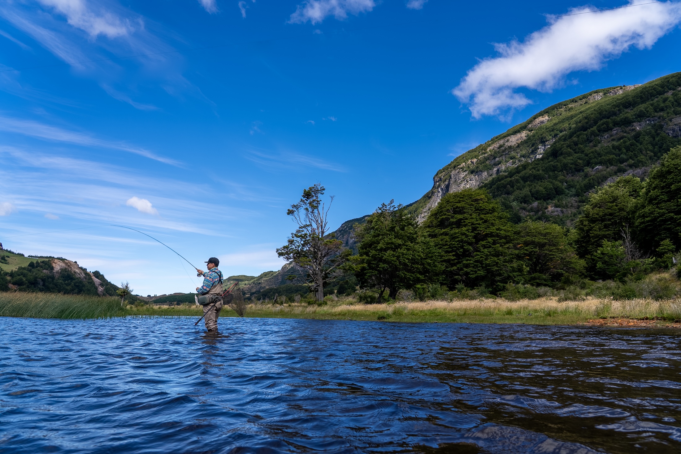 A person standing mid-stream in hipwaders casts a fishing rod