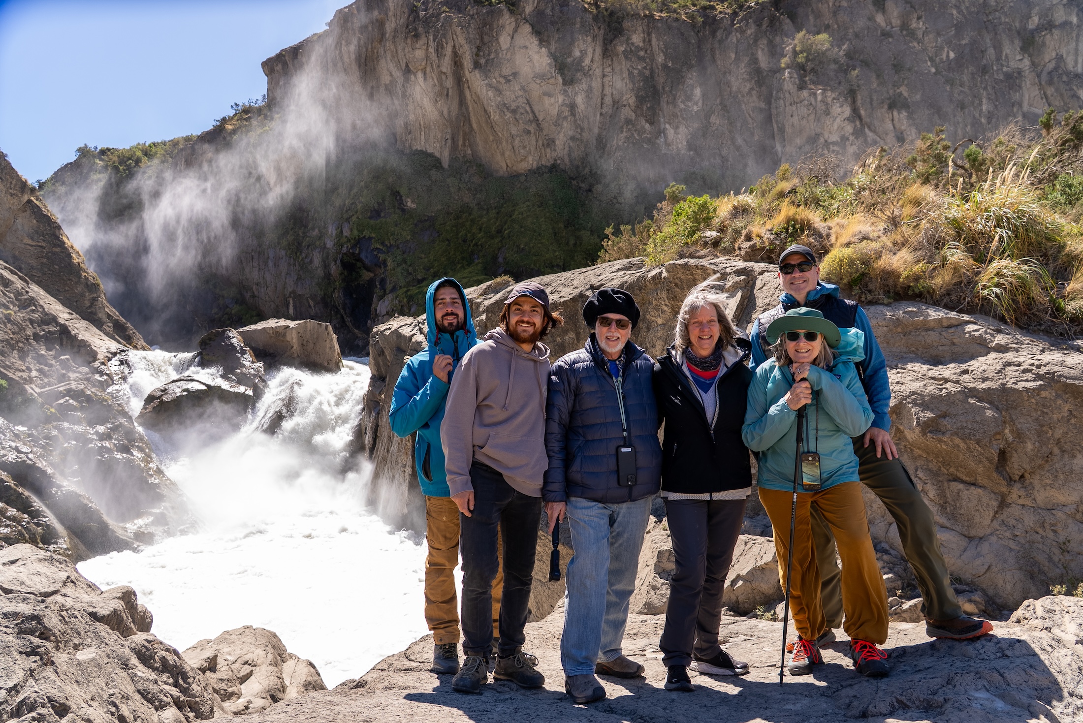A group of six smiling people pose for a photo, a rushing rapid through mountains on their right