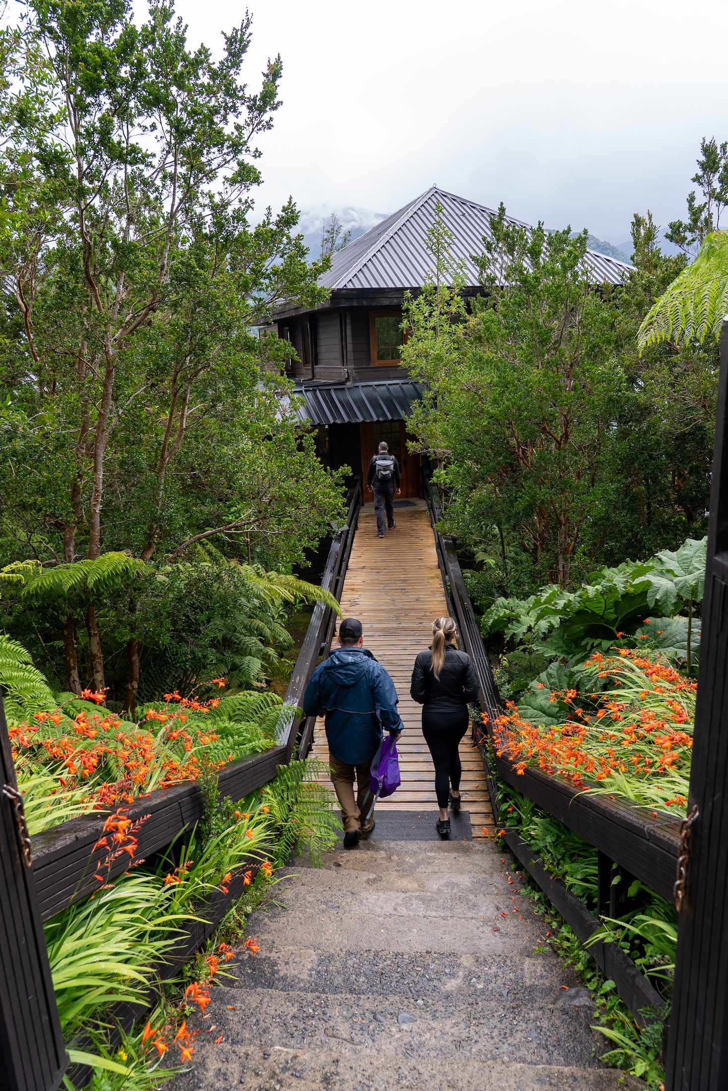 Three people walk down steps with gardens on either side towards a cedar lodge below