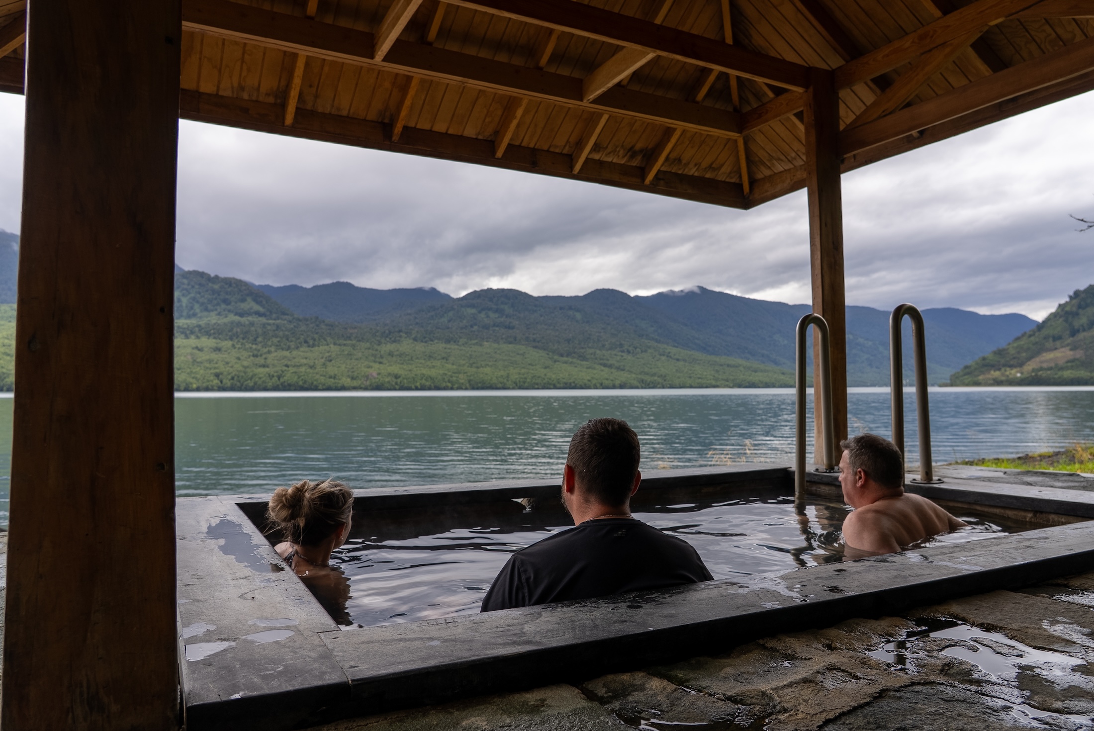 Three people in a hot tub look across the nearby lake towards mountains in the distance