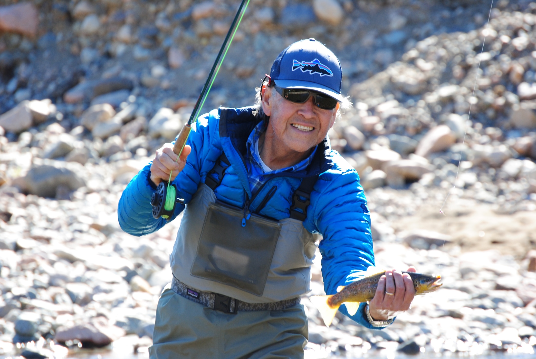 A grinning fisherman in a ballcap holds a fish towards the camera