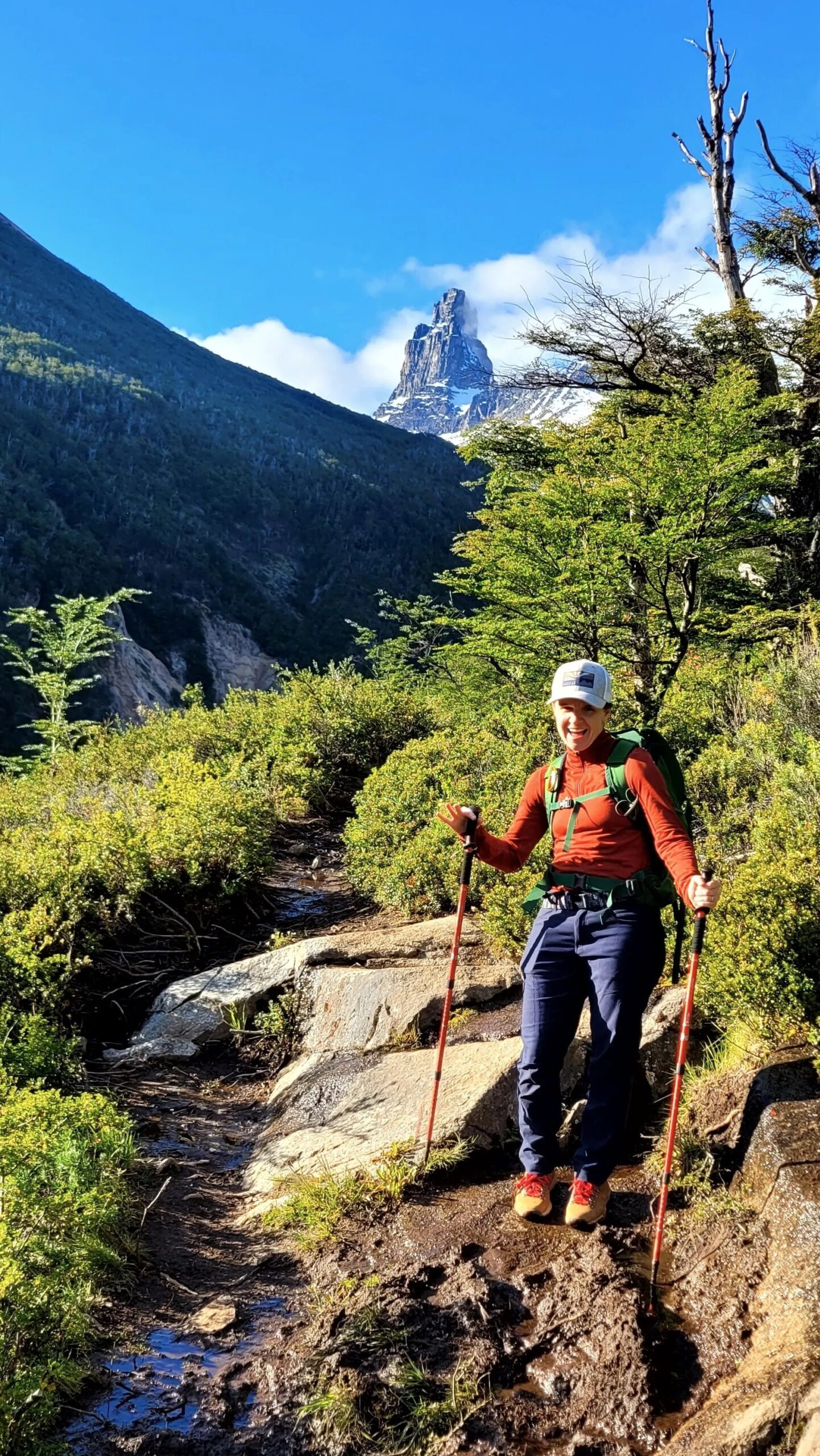 A smiling person in an orange jacket with hiking poles pauses on a trail; a snow-capped mountain is seen in the distance