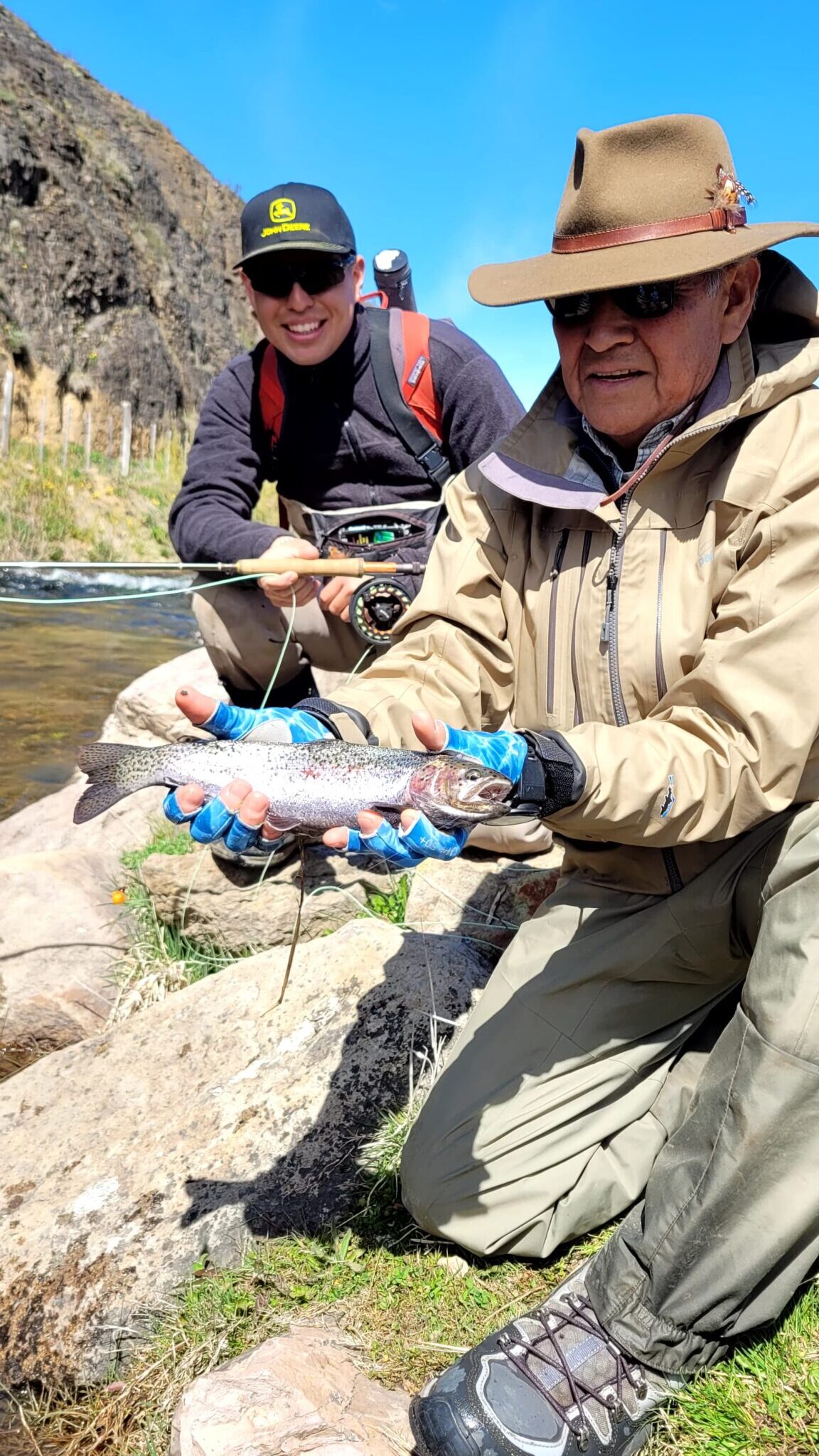 A man in a broad-brimmed hat holds a silver fish in his palms towards the camera; a smiling man squats behind him holding a fishing rod